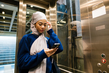 Muslim woman standing in modern elevator using smartphone