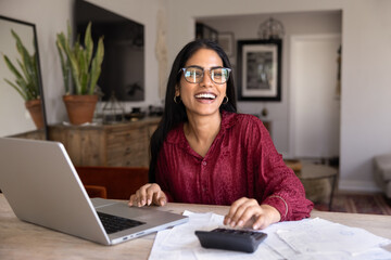 Cheerful young adult Indian girl in glasses counting income and expenses, enjoying management of...