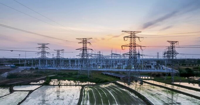 Aerial time-lapse of a large substation at dusk, power infrastructure amid golden-hour glow and reflective rice fields.