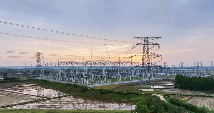 Aerial time-lapse of a large power substation at dawn, with high-voltage towers and transmission lines silhouetted against a soft morning sky over rural farmland.  