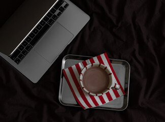 A cup of cocoa on a metal tray, a laptop on brown linens, top view. Lazy morning