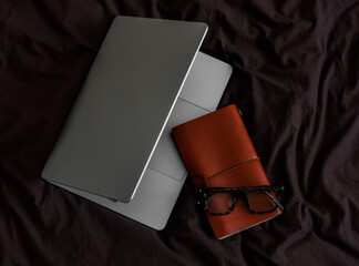 Laptop, leather cover notebook, glasses on brown bed linen
