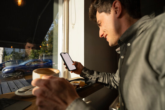 Young Man Enjoys Coffee and a Sandwich While Using Phone