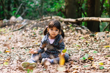 A little girl is sitting on the ground in a forest. She is wearing a black and gray jacket and has her hair in pigtails