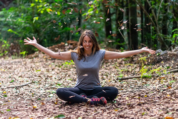 A woman is sitting on the ground and smiling. She is surrounded by leaves and is holding her arms out