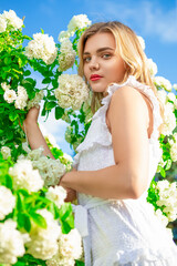 Cute Airy Light Caucasian Fashion Blond Girl in Fragrant Pink Flowers Posing in White Dress in Summer Blossom Park