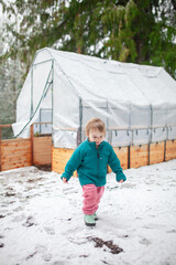 Toddler enjoys free outdoor play in the winter snow beside a backyard greenhouse garden bed. Outdoor parenting, forest school, slow living, family life, and year-round gardening in cold climates.