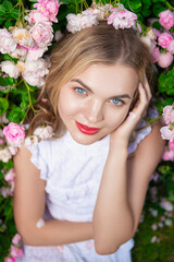 Closeup of Winsome Caucasian Fashion Blond Girl in Fragrant Pink Flowers Posing in White Dress Sitting on Grass in Summer Blossom Park as Fashion, Flowering, Gardening, Cosmetics Concepts.