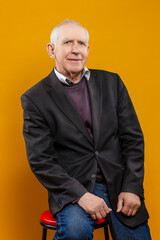 Elderly People Lifestyle Concepts. Portrait of Happy Positive Elderly Gray-Haired Caucasian Man 70s Years Old Wears Brown Jacket and Shirt Looking Straight Smiling Isolated on Yellow Background