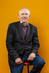Elderly People Lifestyle Concepts. Portrait of Happy Positive Elderly Gray-Haired Caucasian Man 70s Years Old Wears Brown Jacket and Shirt Looking Straight Smiling Isolated on Yellow Background