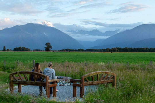 Two caucasian ladies sitting on wooden bench enjoying view of mountains while drinking wine.