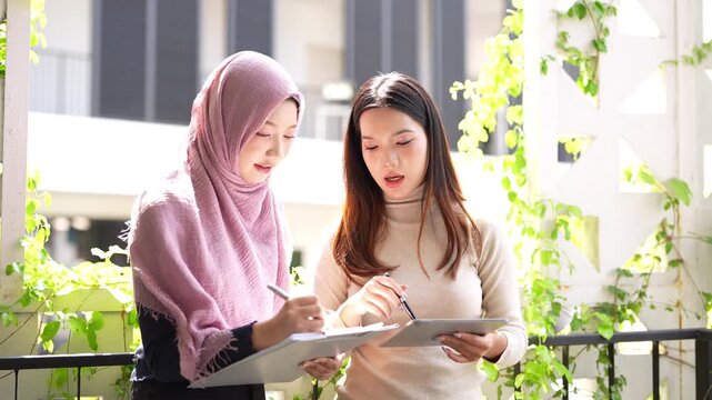 Asian business professional woman talking to Muslim female coworker in hijab, discussing cooperation, teamwork, Standing at corridoor, smiling, giving consultation, advice.