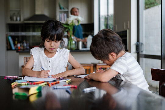 Two kids are busy drawing at a kitchen table, while an adult stands in the background