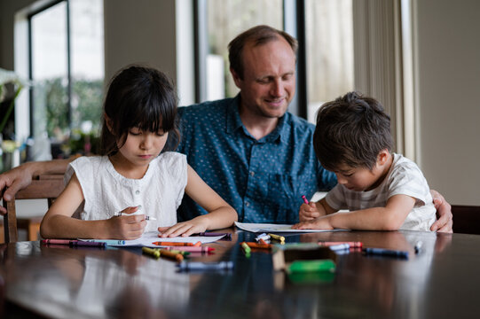 Two children are drawing with crayons at a table while their father assists them with smiles