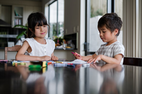 Two children engaged in creative drawing activities at a dining table in a sunny room