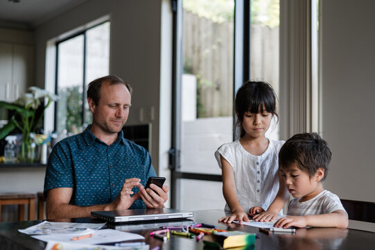 A father uses his phone as two young children draw with crayons at a table