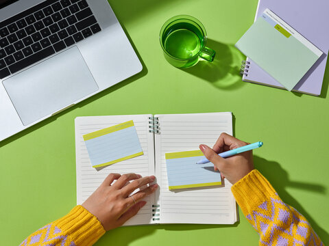 Woman writing notes on a green desk with a laptop and a glass of water