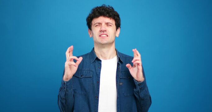 Handsome man with crossed fingers looking at camera having wish and dream on blue background