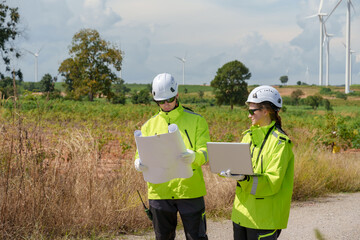 Caucasian adult engineer worker reviewing blueprint and laptop together during wind farm inspection, safety uniform field work supporting wind turbine installation and renewable electricity generation