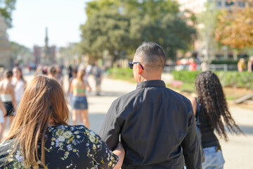 Naklejka premium A man in a black shirt is walking with two women. The man is wearing sunglasses and the women are wearing skirts. The scene is lively and bustling with people walking around