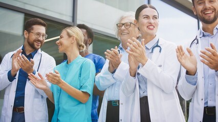 Smiling medical staff clapping hands while standing outside clinic under bright daylight. Confident group celebrating progress and sharing uplifting energy. Professionals enjoying supportive moment.