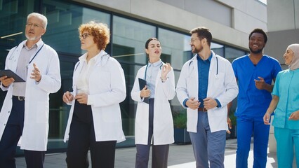 Focused doctors walking outside clinic discussing medical tasks while carrying tablets. Confident colleagues sharing thoughts and reviewing plans during movement. Coordinating next steps together.