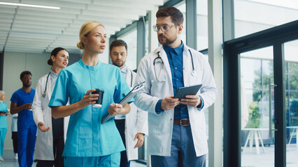 Medical staff walking through clinic hallway enjoying short break. Nurse holding coffee and clipboard talking with male doctor reading tablet. Team moving forward sharing relaxed moment together.