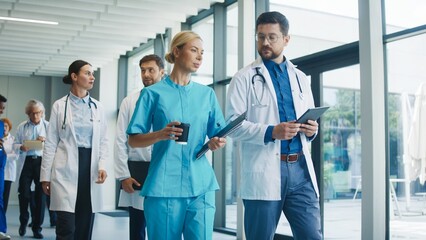Medical staff walking through clinic hallway enjoying short break. Nurse holding coffee and clipboard talking with male doctor reading tablet. Team moving forward sharing relaxed moment together.