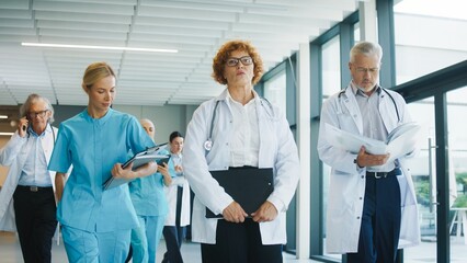 Focused doctor walking through clinic hallway holding clipboard with calm expression. Confident female carrying documents and leading team. Senior male reviewing papers while preparing task.