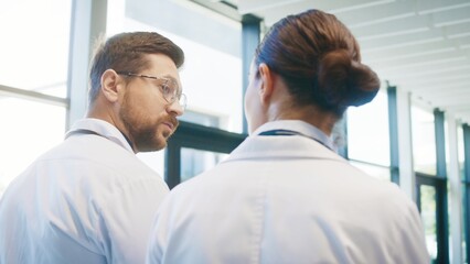 Close camera view from behind two focused doctors walking through bright clinic hallway. Professionals talking casually while moving toward duties. Team preparing for busy day together.
