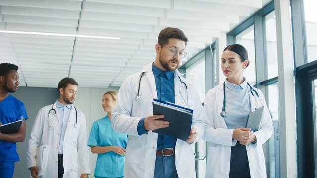 Medical staff walking through bright clinic hallway talking with each other. Focused male doctor reviewing notes while colleague discuss plan. Nurse joining conversation and sharing thoughts.