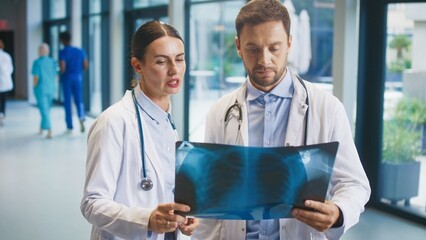 Two focused doctors standing in bright clinic hallway studying chest X-ray together. Female colleague pointing at detail while male doctor examining image closely. Professionals reviewing findings.