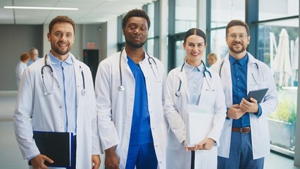 Smiling medical team standing in bright hospital corridor. Male doctor holding folder. Colleagues staying confident. Female doctor standing with documents. Another male doctor holding tablet.
