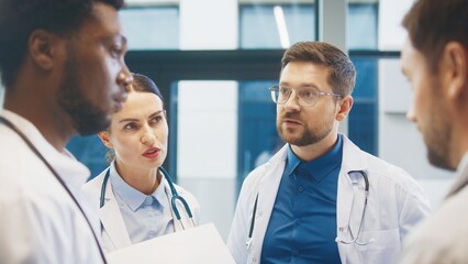 Focused doctor standing with colleagues in bright hospital room. Discussing patient update. Sharing notes and coordinating next steps during calm professional exchange. Relaxing during lunch break.