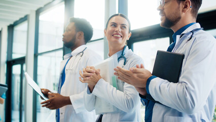 Cute female doctor smiling and clapping in bright hospital corridor. Male colleagues joining applause. Team celebrating success and appreciating strong performance during busy shift.