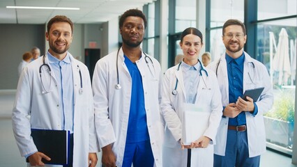 Smiling medical team standing in bright hospital corridor. Male doctor holding folder. Colleagues staying confident. Female doctor standing with documents. Another male doctor holding tablet.