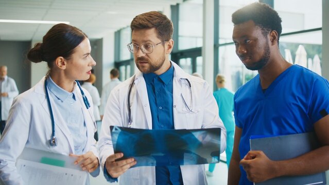 Male doctor examining chest X-ray while discussing treatment options with colleague in bright medical corridor. Focused expression. Evaluating condition for correct prescription. Healthcare concept. - Powered by Adobe