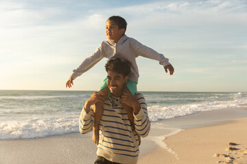 Happy father carrying son on shoulders with arms outstretched enjoying sunset at beach