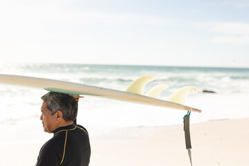 senior man carrying surfboard over head at beach against sky during sunny day