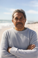 Portrait of retired senior man with arms crossed at beach against sky during sunny day