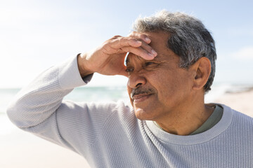 Retired senior man looking away while shielding eyes with hand at beach on sunny day