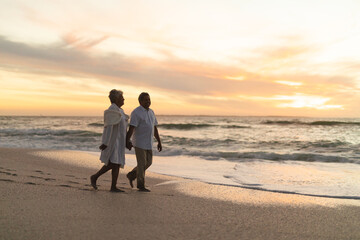 Senior multiracial couple enjoys sunset beach walk against sky