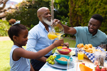 Smiling african american man serving juice for daughter by senior man at dining table in backyard