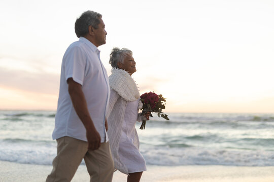 Side view of newlywed senior man and woman with bouquet walking at beach during sunset