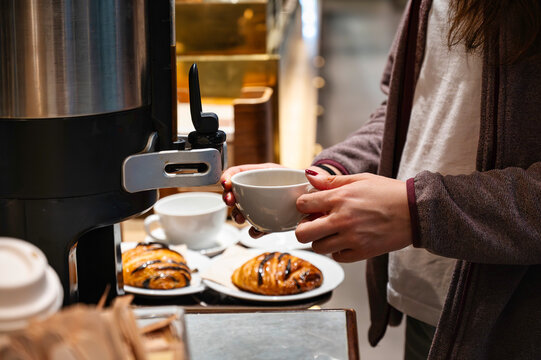 Woman holding white cup at coffee dispenser in self-service breakfast area with croissants and beverages in modern caf&eacute; or airport environment