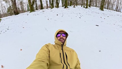 Young man with sunglasses recording a selfie video while talking and walking backwards through a snowy forest, eventually falling into the fresh snow creating a funny moment