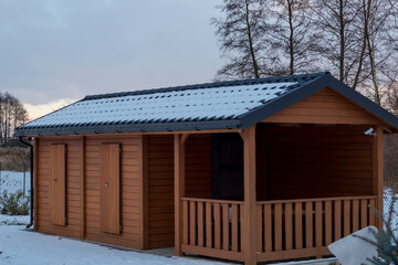 Drewniany domek położony na działce rekreacyjnej, pod zachmurzonym,zimowym niebem otoczony spadłym lekkim śniegiem. Budynek ma skośny, ciemny, metalowy dach pokryty warstwą śniegu. 