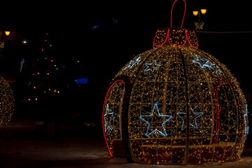 Dekoracje świąteczne na gł&oacute;wnym placu miasta. Duża, podświetlana konstrukcja bombki ozdobiona gwiazdkami w kolorze niebieskim i ciepłej bieli, osadzona w nocnej scenerii miasta. Czerwone światełka .