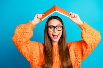 Young woman with orange sweater holds book over her head smiling against a blue background