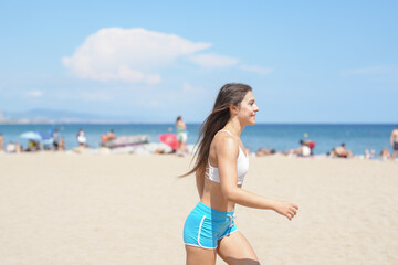 Fototapeta premium A woman is walking on the beach wearing a white top and blue shorts. She is smiling and she is enjoying her time. The beach is crowded with people, some of whom are sitting on the sand